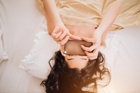 Beautiful girl with black wavy hair naked taking a selfie on phone lying in the morning in bed. Indoor shot of cheerful caucasian woman has morning video call, holds modern cell phone.の写真素材