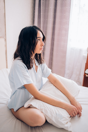 Beautiful young woman with dark hair wearing nightwear sitting on bed in cozy bedroom having pleased smile looking away, remembering an unforgettable night with her boyfriend. Dreamy, relaxed girl.の写真素材