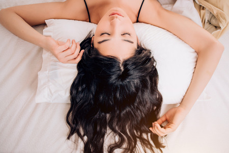 Close up face of young beautiful black wavy haired woman lying on white pillow with closed eyes, hands over head, view from above. Hair care, relaxation, sleep concept, crop photoの写真素材