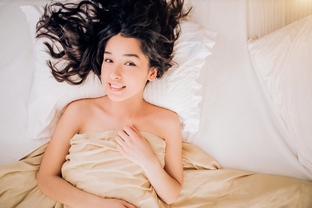 Romantic young beautiful female model with happy expression and naked body covered with blanket poses on white bedclothes in comfortable bed at hotel room, enjoys starting new day. Top view.Copy spaceの写真素材