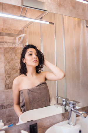Beautiful brunette woman in bathroom looking in mirrorの写真素材