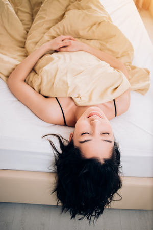 Good morning. Relaxed nice brunette lies on soft bed at home, enjoys with closed eyes after healthy dream, has good mood in morning. Positive emotions, good sleep, relaxation concept.の写真素材