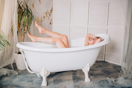 Total relaxation, purity, sensuality. Beautiful young caucasian with perfect soft skin showing her slender bare legs while lying in bathtub in modern spacious bathroom with green plants.の写真素材