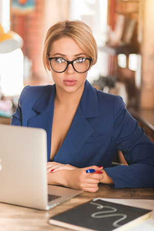 Closeup portrait of beautiful business lady with blonde hair, plump lips, makeup, wearing formal wear, in big stylish glasses sitting in front of computer looking at camera while working in office.の写真素材