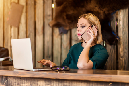 Caucasian woman with short haircut dressed in green sweater having conversation by smartphone with client while sitting at her workplace looking at laptop with smile. Business and technology concept.の写真素材