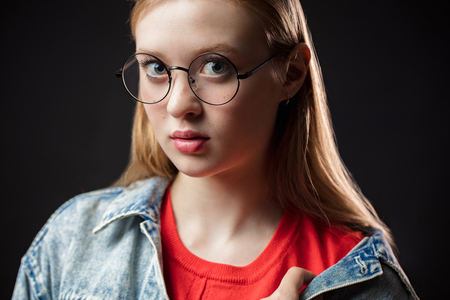 Close up portrait of a young redhaired woman in glasses on a black background. The girl looks at the camera. Emotional face, expressive facial featuresの写真素材