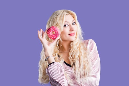 Joyful caucasian blonde woman having delicious pastry, holding pink sweet glazed doughnut, enjoying tasty dessert isolated against purple blue background. Holiday, unhealthy food, nutrition concept.の写真素材