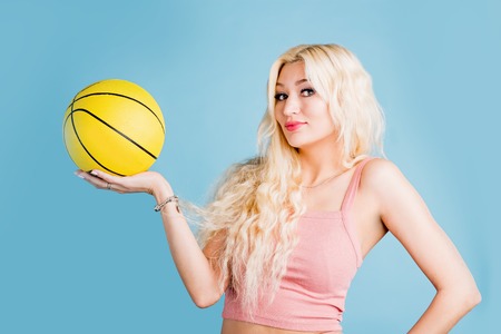 Sporty girl in a pink short t shirt holding a yellow basketball smiling looking at camera on light...