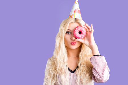 Surprised blonde long haired lady wearing dress and party hat covering face with tasty pink doughnuts, having amazed expression, isolated over purple studio wall. Delicious snack, birthday, holiday.の写真素材
