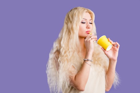 Attractive young caucasian woman with long blonde hair, holding takeaway yellow paper cup, drinking soda with straw, enjoying tasty beverage with closed eyes , posing against violet background.の写真素材