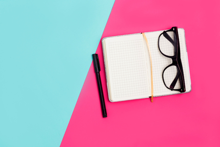 Flat lay, top view office table desk. Work space with open Notepad, glasses and pens on pink and blue paper background with copy spaceの写真素材