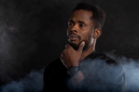 Headshot of serious dark skinned male holding chin, dressed in black windbreaker jacket, wristwatces, standing against dark background with white smoke. Close up.の写真素材