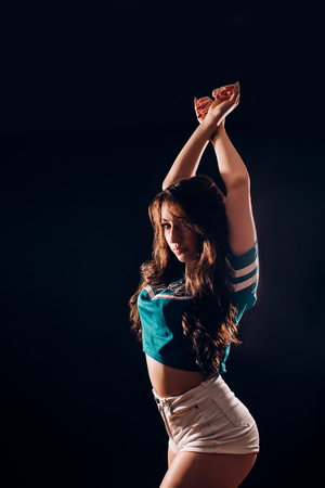 Portrait of sexy attractive hot brunette woman with long wavy hair in short white shorts and blue t-shirt in the dark on black background. Slim figure, fitness, sports, dancing, twerk, hobby conceptsの写真素材