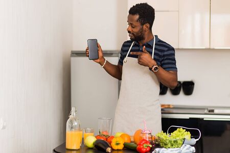 Cooking, technology, advertising concept. Portrait of happy handsome african american young man in apron holding smartphone with blank black screen and pointing it with finger.の写真素材