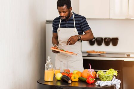Handsome young bearded african american man cooking at home kitchen, cutting cucumbers and tomatoes, preparing breakfast for girlfriend. Healthy food concept.の写真素材