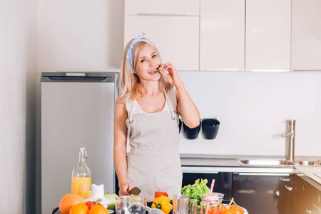 Positive young woman posing in kitchen at home. Blonde girl wearing apron, holding knife, preparing vegetable salad, eating cucumber, smiling looking at camera . Healthy food, lifestyle concept.の写真素材