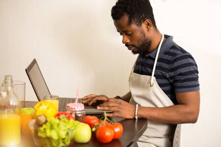 Young bearded african man wearing apron working on laptop, sitting at table in the kitchen, while preparing dinner, drinking fresh fruit juice. Side view photo. Cooking and technology concept.の写真素材