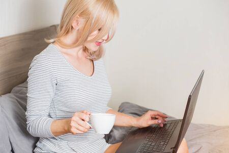 Beautiful caucasian blonde girl in sleepwear having cup of coffee, looking at screen of laptop sitting in bed in bedroom at home. Side view photo. People, leisure, technology, freelance concept.の写真素材