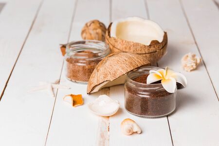 Close-up of chocolate coconut scrub for face and body in small jars with shells and a flower on a white wooden background. Spa, body care, sea, relaxation concept.の写真素材
