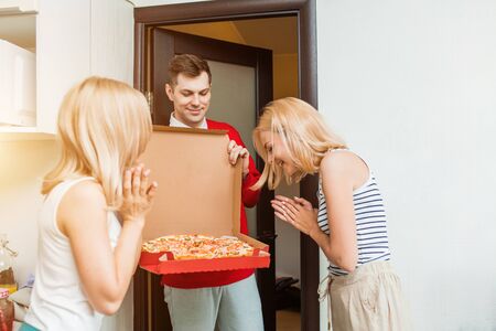 Two happy young caucasian women receiving pizza from delivery man at home. Delivery man in red uniform showing pizza to customers at the door. Food Delivery.の写真素材