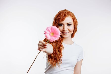 Young beautiful woman with pink flower on white background with copy space. Emotional caucasian redhead girl laughing to camera and gives a pink gerbera.の写真素材