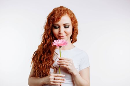 Close up portrait of pretty caucasian woman smelling a flower. Cute young girl with curly ginger hair, fresh skin holding one flower over white background with copy space and looking down.の写真素材