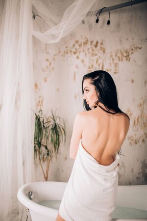 Backside portrait of slim attractive brunette woman covering her body with white towel standing near bathtub looking to side in modern bathroom. Sensuality, purity, and beauty concept.の写真素材