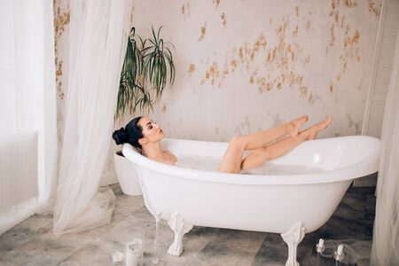 Beautiful young woman with black hair bun, perfect skin showing her slender bare legs while lying in bathtub in modern luxury bathroom with green plants, candles. Total relaxation, purity, sensuality.の写真素材