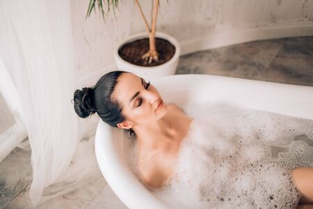 Sensual sexy woman with black hair bun, pretty face with makeup, lying in bath with closed eyes, enjoying good rest, relaxing in foam water with in bathroom. Pleasure, Spa and beauty concept. Top view.の写真素材