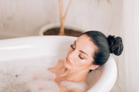 Indoor shot of female model lying in bath. Closeup portrait of beautiful relaxed young black haired...