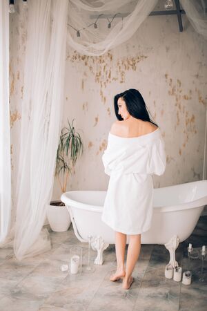 Rear shot of brunette spa caucasian girl with black long hair, wearing white bathrobe, preparing to...
