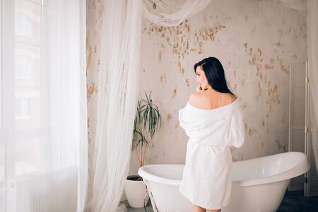 Young beautiful caucasian elegant woman with long dark hair wearing white bathrobe preparing to take a bath in loft interior bathroom. Morning wellness hygiene procedures.の写真素材