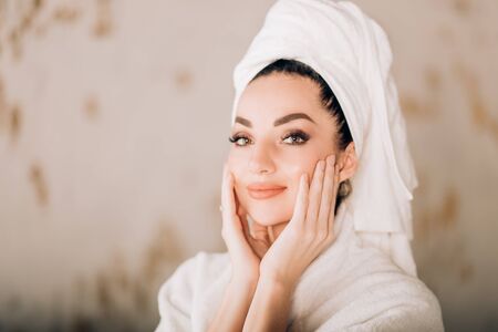 Closeup shot of young beautiful woman applying moisturizing cream on her face, touching her face enjoying soft fresh skin after spa procedures looking with cute smile at camera. Beauty and spa conceptの写真素材