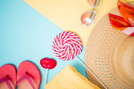 Beige straw beach sun hat, towel, pink flip flops and red lollipops on pastel sky blue yellow background.の写真素材
