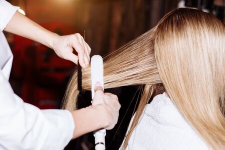 Close up of process straightening long blonde hair with hair irons. Beauty, hairstyle, hot styling,...