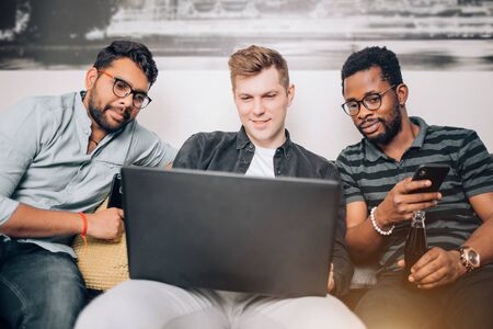 Group of cheerful male friends students sitting together on sofa at home and watching funny video on laptop, playing computer games, using mobile phone. Friendship, technology, entertainment concept.の写真素材