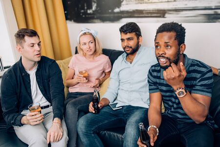Group of multi ethnic friends having fun at home, talking , enjoying drinks sitting on couch in...