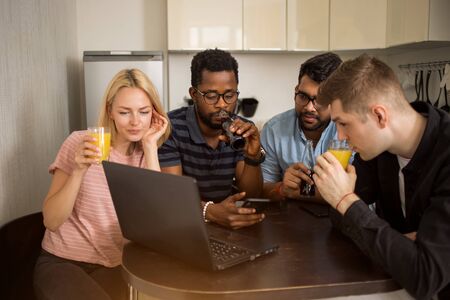 Group of diverse students sitting at table, drinking juice, using mobile phone and laptop, watching video together. Four friends having fun in kitchen. Technology, communication concept.の写真素材
