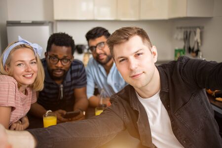 Say Cheese. Handsome caucasian man taking selfie with friends. Happy multi ethnic students sitting at table and having lunch in kitchen at home.の写真素材