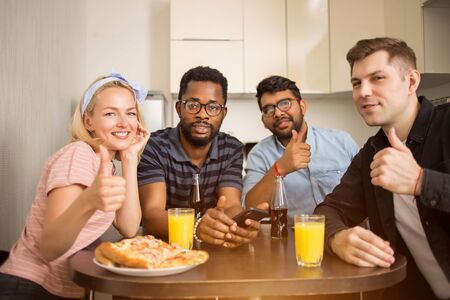 Group of diverse friends sitting at table in kitchen showing thumb up gesture, eating tasty pizza, drinking juice and beer, smiling looking at camera. People, entertainment, food delivery concept.の写真素材