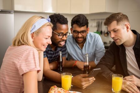Beautiful happy young caucasian blonde woman sitting at table with her friends, laughing looking at smartphone. Multi ethnic students testing new gadget. Technology, friendship, leisure concept.の写真素材