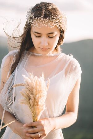 Beautiful girl bride in a dress holding a bouquet of grasses, the delicate decoration on the head,...