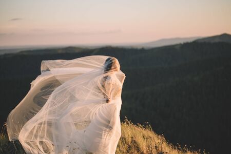 Portrait of a beautiful young brunette bride woman in a white dress and with a long veil Curling in the wind in nature, the mountains in summer. Fine art, wedding, lifestyle, love, life conceptの写真素材