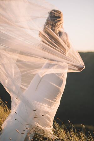 Slender woman in a long white dress and with a veil Curling in the wind, view from the back, outdoors in summer. Beautiful transparent fabric, tulle, satinの写真素材