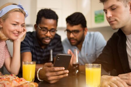 Four friends sitting at table, eating pizza, drinking soft beverages, reading text message on smartphone, testing new phone application together. Multicultural students having fun in hostel kitchen.の写真素材
