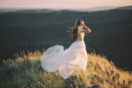 Brunette woman in white wavy dress in the wind in the mountains, side view. Fine art, wedding, happiness, lifestyle, love, life conceptの写真素材