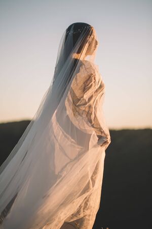 Sideways portrait of the bride in a long white dress in nature summer sunset