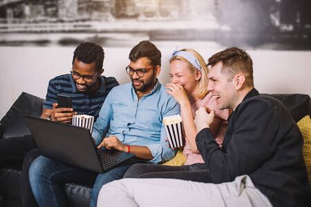 Cheerful diverse friends students sitting together on sofa at home and watching funny video on...