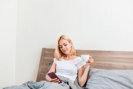 Relaxed european girl in white t shirt drinking coffee sitting on cozy bed. Indoor portrait of good...