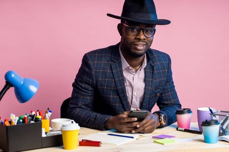 Happy stylish handsome afro american businessman sitting at desk surrounded with stationery, wearing...